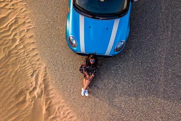 Mini Cooper azul segunda mano Foto aérea de un Mini cooper azul aparcado en la playa con una mujer sentada a su lado
